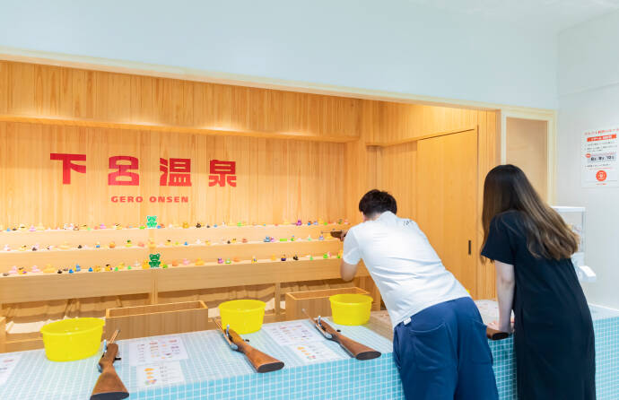 射的場で射的を楽しむ男女。男性が射的用の銃を構えて狙いを定めている。女性が後方で見守っている